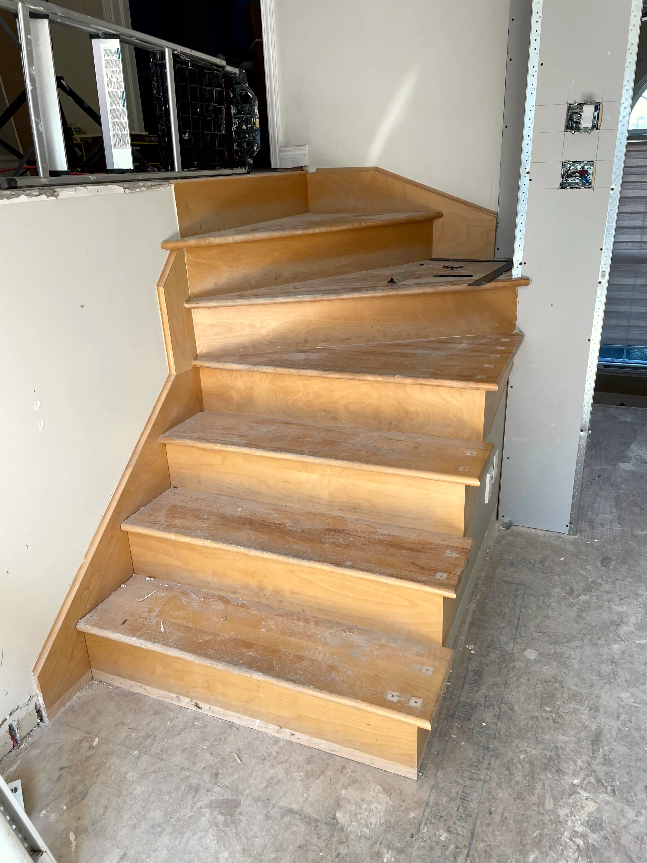 New plywood staircase with winder treads against a drywalled wall fitted with an electrical box.