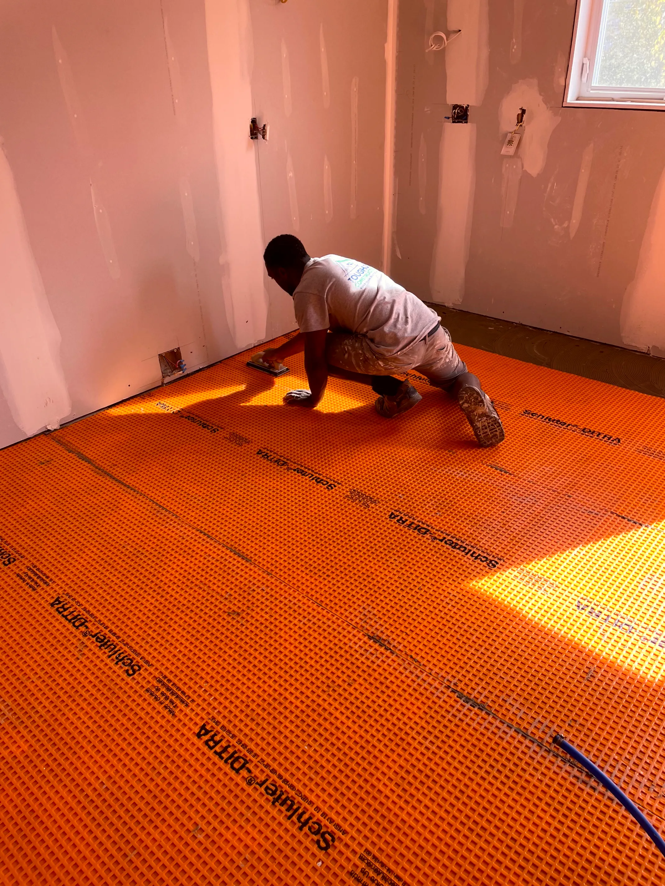 Worker troweling an orange floor membrane across a drywalled room with a corner window.