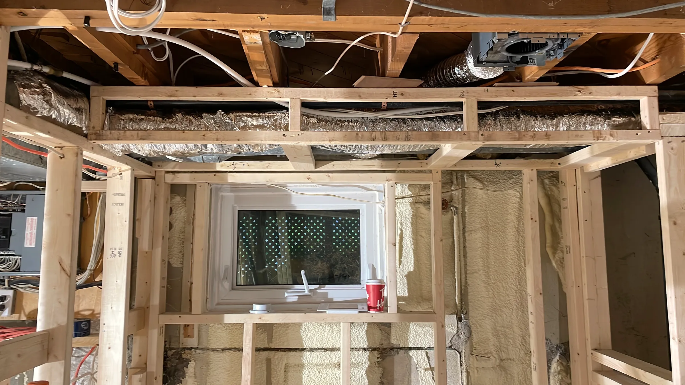 Basement framing with a soffit, ductwork, and wiring overhead around a small window.