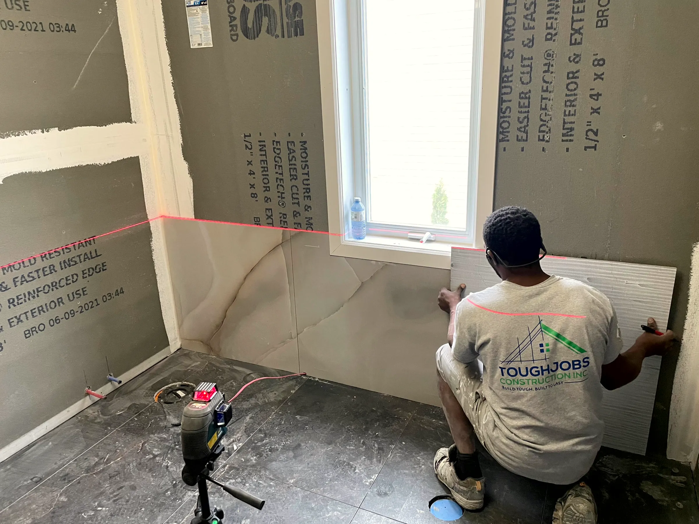 Worker setting bathroom wall board to a laser line on cement board beside the window.