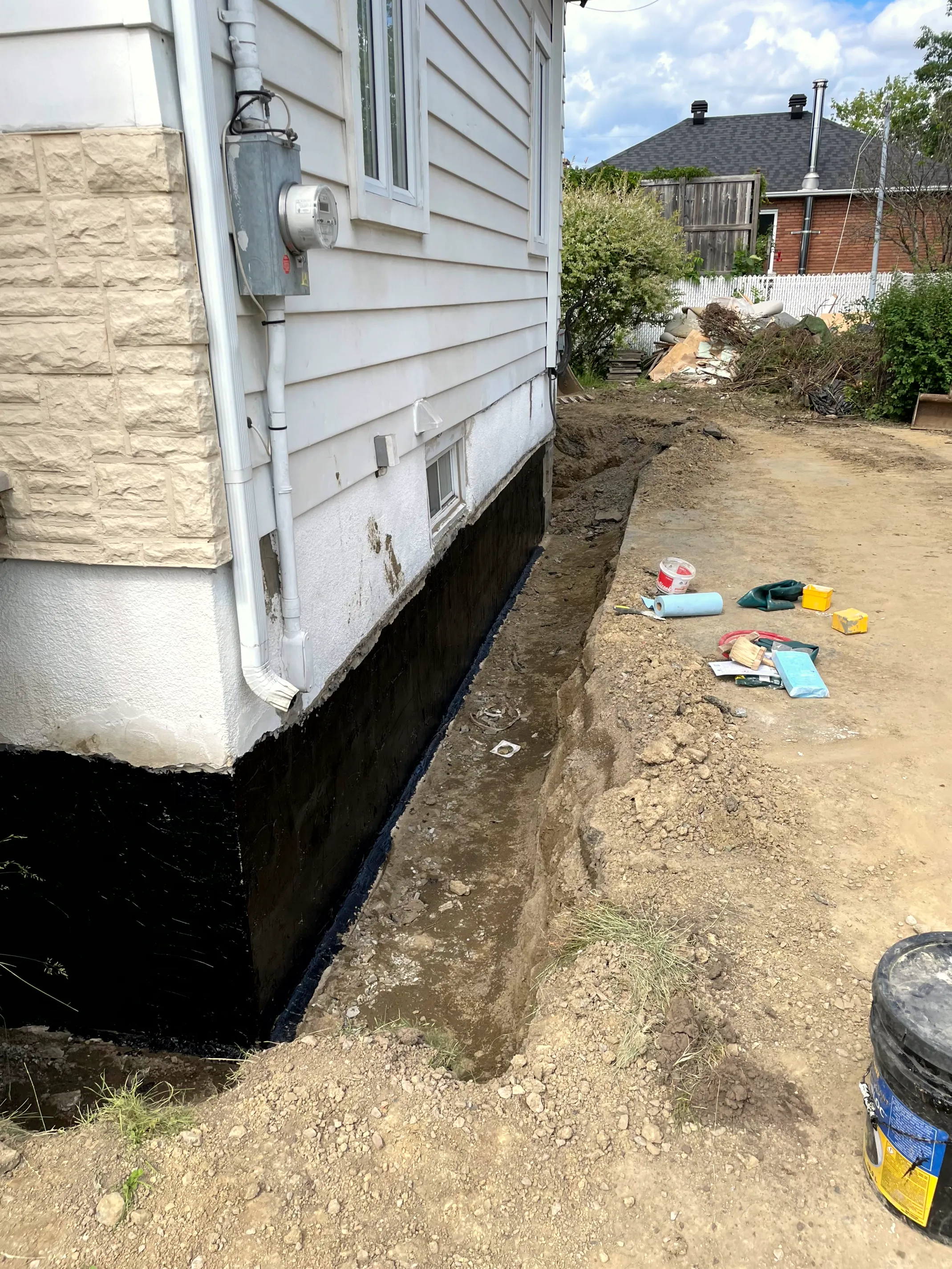 Exterior foundation wall coated with black waterproofing along an excavated trench beside the house.
